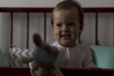 Gri bebek beşiğinde oturan sevimli, neşeli, tombul bir kız bebek. Beyaz ve pembe pijamalarla uyanıp, ayıcık gibi gülümsüyor. Çocukluk, rahatlık, rahatlık, ilaç, sağlık konsepti