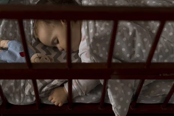 Linda niña regordeta alegre que duerme dulcemente en cuna gris durante el descanso del almuerzo en pijamas blancos y rosados con oso de peluche en casa. Infancia, ocio, comodidad, medicina, concepto de salud — Foto de Stock
