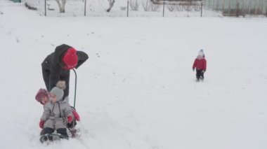 Tatil, oyun, aile kavramı, yavaş çekim otantik üç mutlu anaokulu çocuğu, genç Wonan annesi kızakla kayan ve karla oynayan kardeşleri. Soğuk mevsimde kar yağışı açık havada kış bahçesinde