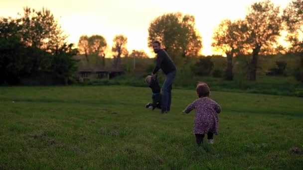 Οι άνθρωποι στο πάρκο. χαρούμενη οικογενειακή σιλουέτα το ηλιοβασίλεμα. Ο μπαμπάς κουνάει το μωρό απ 'τα χέρια του και περνάει χρόνο. γονείς και διασκεδαστικά παιδιά που περπατούν σε ανοιχτό χώρο. Ημέρα πατέρων, παιδική ηλικία, έννοια της πατρότητας — Αρχείο Βίντεο