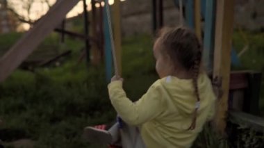 Sarı gri salıncağa binip bahar günbatımında çimenlere düşen otantik şirin anaokulu kızı. Güneş doğarken doğadaki çocuk. Çocukluk, ebeveynlik, aile, yaşam tarzı konsepti