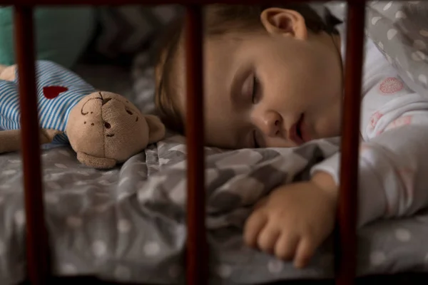 Linda niña regordeta alegre que duerme dulcemente en cuna gris durante el descanso del almuerzo en pijamas blancos y rosados con oso de peluche en casa. Infancia, ocio, comodidad, medicina, concepto de salud — Foto de Stock