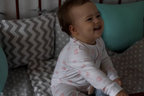 Bonito alegre pequena menina gordinha sentada no berço do bebê cinza depois de acordar do sono em pijama branco e rosa com ursinho de pelúcia e sorrindo. Infância, lazer, conforto, medicina, conceito de saúde — Fotografia de Stock