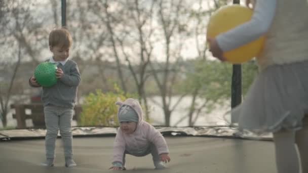 Primavera, deporte, día de fiesta, familia, infancia - cámara lenta Tres hermanos preescolares niños pequeños felices niños gemelos jugar divertirse saltar bolas en trampolín al aire libre soleado atardecer en el parque — Vídeo de stock