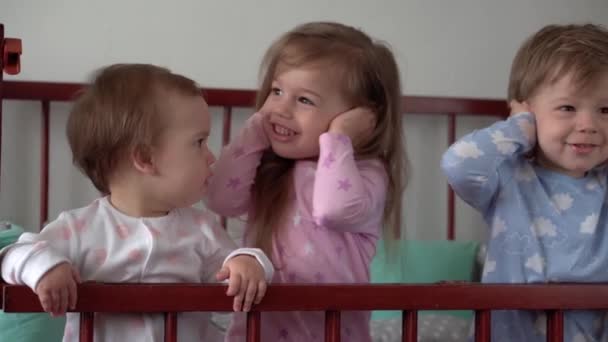 Cute cheerful 3 little kids girl and boy siblings preschool children in grey baby crib after waking up from sleep in pajamas with legs dangling. Childhood, leisure, comfort, medicine, health concept — Stock Video