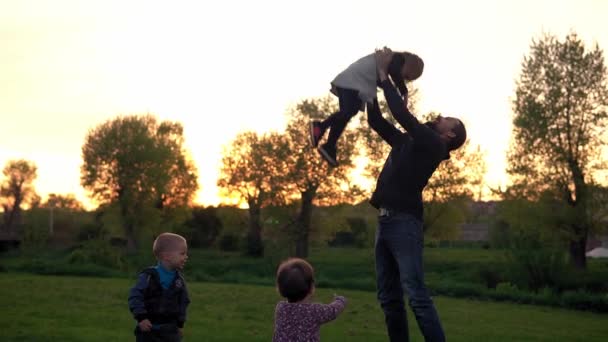 公園の人たち。日没時の幸せな家族のシルエット。空気中の子供を投げるお父さんは時間を費やしています。親と楽しい子供たちが野外で歩いています。父の日、子供の頃、親の概念 — ストック動画