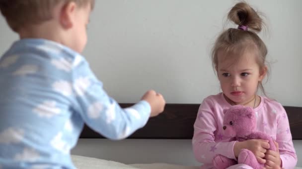 2 leende förskolebarn pojke och flicka leka med nalle i blå rosa pyjamas på sängen. Små tvillingar har kul, glada barn i karantän hemma. Vänskap, familj, utbildningskoncept — Stockvideo