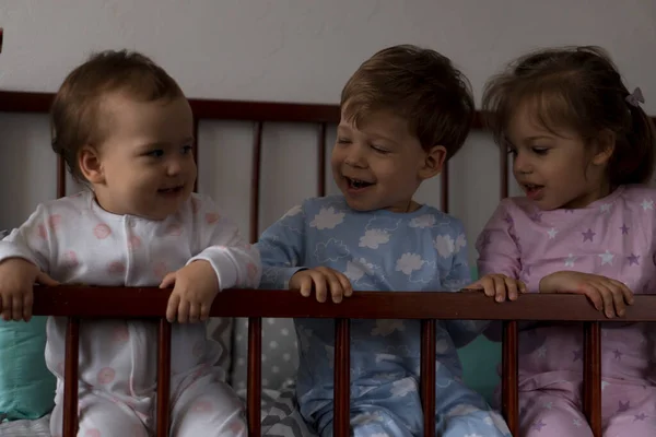 Cute wesoły 3 małe dzieci dziewczynka i chłopiec rodzeństwo przedszkolne dzieci w szarości łóżeczko po przebudzeniu ze snu w piżamie z nogami zwisające. Dzieciństwo, czas wolny, komfort, medycyna, koncepcja zdrowia — Zdjęcie stockowe