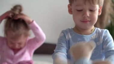 İki tane gülümseyen anaokulu çocuğu erkek ve kız çocuğu yatakta mavi pijamalı oyuncak ayıyla oynuyorlar. Küçük bebek ikizler evde karantinada mutlu çocuklar. Arkadaşlık, aile, eğitim kavramı