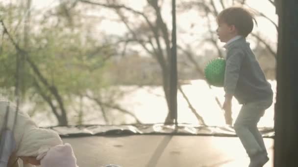 Frühling, Sport, Urlaub, Familie, Kindheit - Zeitlupe Drei glückliche kleine Kleinkinder Vorschulgeschwister Kinder Zwillinge Kinder spielen haben Spaß beim Bällchenspringen auf dem Trampolin draußen sonniger Abendsonnenuntergang im Park — Stockvideo