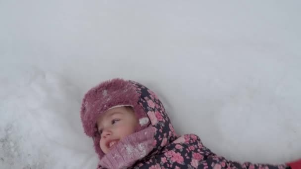 Vakantie, spel, kindertijd concept - slo-mo authentieke vrolijke kleuter peuter baby meisje glimlach ligt in de sneeuw op de rug en applaus met voeten maken engel. sneeuwval in het koude seizoen weer in de winter buiten — Stockvideo