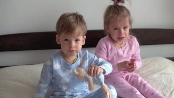 2 niños en edad preescolar sonrientes niño y niña peleando por un juguete en pijama rosa azul en la cama. Los bebés gemelos se divierten, niños felices en cuarentena en casa. Amistad, familia, concepto educativo — Vídeo de stock