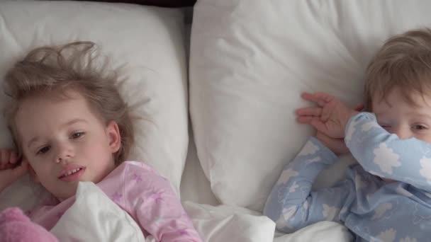 Authentieke Kaukasische 2 kleine broertjes en zusjes peuters peuter meisje jongen in roze blauw slaperig bij het wakker worden kijkend naar camera lachend schattig en grimmig in wit bed. Kinderopvang, Kindertijd, Ouderschap, Levensstijl — Stockvideo