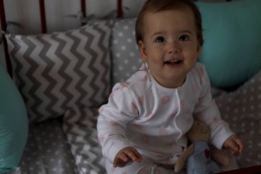 Gri bebek beşiğinde oturan sevimli, neşeli, tombul bir kız bebek. Beyaz ve pembe pijamalarla uyanıp, ayıcık gibi gülümsüyor. Çocukluk, rahatlık, rahatlık, ilaç, sağlık konsepti