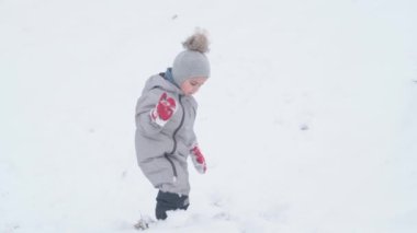 Tatil, oyun, çocukluk konsepti - yavaş çekim otantik mutlu doğu anaokulu çocuğu bebek gülümsemesi kameraya bakıyor ve derin karda yürümeye çalışıyor. Soğuk mevsimde kar yağışı Kışın açık havada