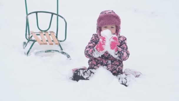 Semester, spel, barndomskoncept - and mo äkta glad förskola småbarn flicka leende sitter håller hög med snö i händerna och äter glatt. snöfall under kalla årstider väder på vintern utomhus — Stockvideo