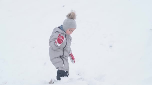 Tatil, oyun, çocukluk konsepti - yavaş çekim otantik mutlu doğu anaokulu çocuğu bebek gülümsemesi kameraya bakıyor ve derin karda yürümeye çalışıyor. Soğuk mevsimde kar yağışı Kışın açık havada — Stok video