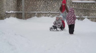Tatil, oyun, aile kavramı, yavaş çekim otantik üç mutlu anaokulu çocuğu, genç Wonan annesi kızakla kayan ve karla oynayan kardeşleri. Soğuk mevsimde kar yağışı açık havada kış bahçesinde