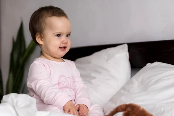 Äkta porträtt kaukasiskt litet spädbarn knubbig liten flicka eller pojke i rosa sömnig när du vaknar tittar på kameran ler söta och grimaser i vit säng. Barnomsorg, Barndom, Föräldraskap, Livskoncept — Stockfoto