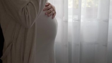Otantik, tatlı, hamile bir kadın evde karnıyla mutlu hissediyor. Genç anne karnını okşuyor ve pencerenin yanındaki camdan su içiyor. Doğum öncesi bakım, hamilelik, annelik, yaşam tarzı konsepti