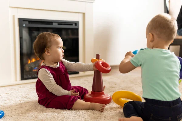 Familia, amigos, amistad, infancia, desarrollo, habilidades, juegos de motilidad - niños pequeños niños auténticos niños pequeños pone anillos en la pirámide jugando juntos con juguete sentarse en el suelo en casa acogedora — Foto de Stock