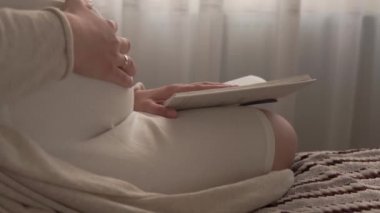 Otantik, tatlı, hamile bir kadın evde karnıyla mutlu hissediyor. Genç anne karnını okşuyor ve pencerenin yanındaki yatakta kitap okuyor. Doğum öncesi bakım, hamilelik, annelik, yaşam tarzı konsepti