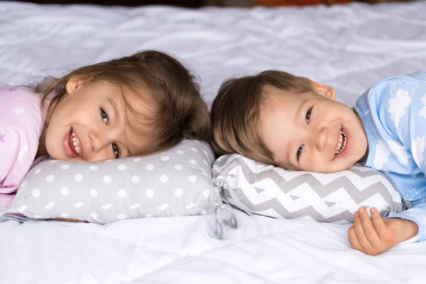 Hogar, comodidad, infancia, cuidado, amistad, dulce sueño- dos sonriendo feliz niño auténtico hermanos niños gemelos en pijama descansar dormir en la cama almohadas mirar a la cámara suave y acogedor estado de ánimo soñoliento — Foto de Stock