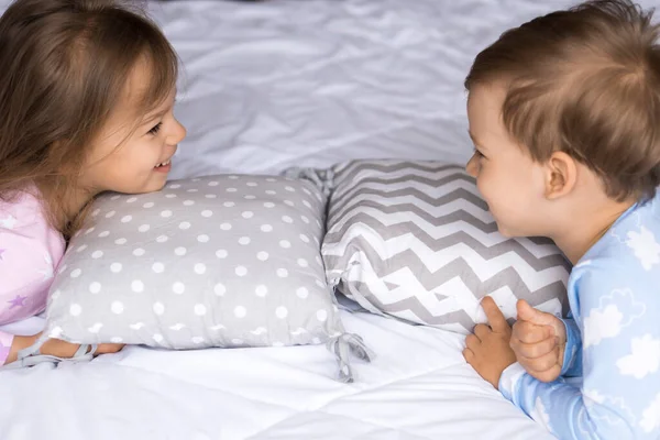 Casa, comfort, infanzia, cura, amicizia - due sorridenti felici autentici bambini fratelli gemelli bambini in pigiama si riposano sui cuscini del letto si guardano negli occhi, morbido accogliente umore assonnato all'interno — Foto Stock