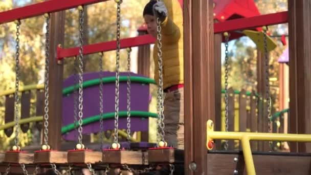 Childhood, family, motherhood, Activities concept - preschool minor child boy 2-4 years in red orange beret playing on playground in cold autumn park. small kid have fun on drawbridge chains outdoors — Stock Video