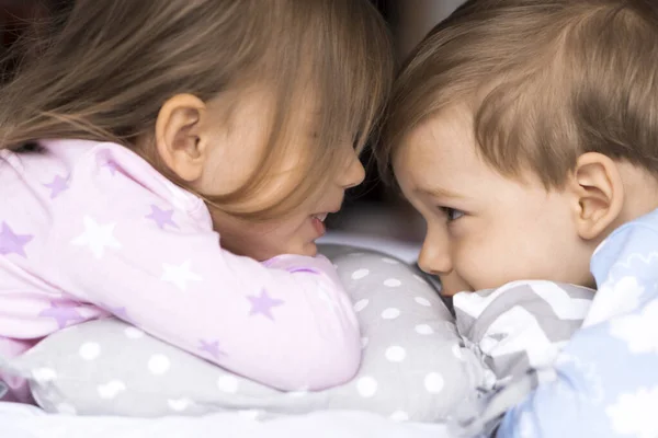 Otthon, kényelem, gyermekkor, gondozás, barátság - két mosolygós boldog autentikus kisgyermek testvér gyerekek ikrek pizsamában feküdni pihenés az ágypárnák néz egymás szemébe, puha hangulatú álmos beltéri — Stock Fotó