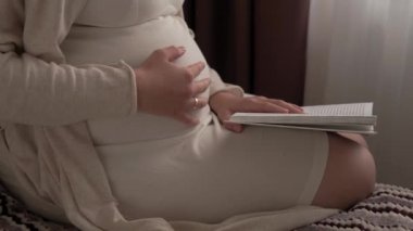 Otantik, tatlı, hamile bir kadın evde karnıyla mutlu hissediyor. Genç anne karnını okşuyor ve pencerenin yanındaki yatakta kitap okuyor. Doğum öncesi bakım, hamilelik, annelik, yaşam tarzı konsepti