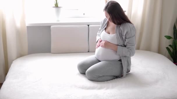 Gravidez, maternidade, pessoas, conceito de expectativa - mulher grávida feliz tocando sua barriga na cama em casa cuidar mãe acaricia grande barriga nua com as mãos, na sala de luz no dia ensolarado de verão — Vídeo de Stock