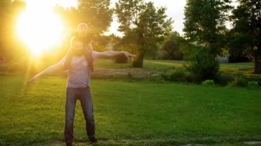 Yaz, tatil, doğa, mutlu aile, çocukluk, babalık, baba günü - küçük bir anaokulu çocuğuyla çocuk çocuk kanat çırpmaca oynuyor çocuklar günbatımında parktaki çayırda omuzlarına oturup eğleniyor.