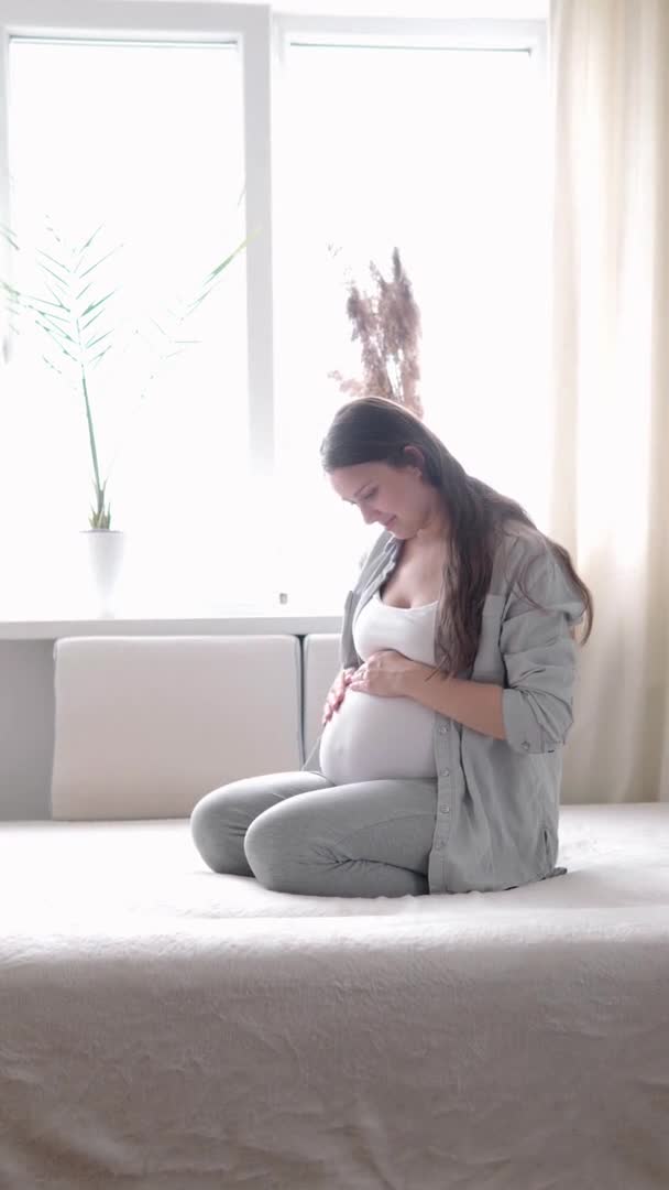 Vertical Format Video de una mujer embarazada feliz tocando la barriga en la cama en casa madre cariñosa acaricia la barriga grande del vientre con las manos en un día soleado de verano. embarazo, maternidad, personas, concepto de expectativa — Vídeo de stock