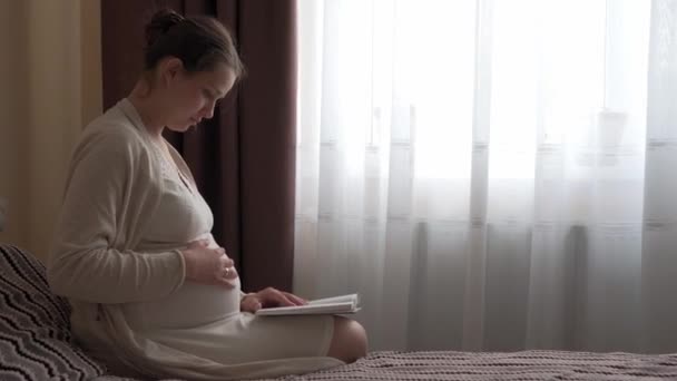 Mulher grávida bonito autêntico sentindo-se feliz em casa com a barriga. Jovem mãe acariciando barriga e lendo livro na cama perto da janela. Maternidade pré-natal, gravidez, maternidade, conceito de estilo de vida — Vídeo de Stock