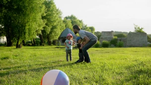 Estate, vacanza, natura, famiglia felice, infanzia, paternità, festa del padre - Papà con bambini piccoli in età prescolare i bambini corrono a giocare si divertono a vomitare per mano con una grande palla gonfiabile nel parco al tramonto — Video Stock