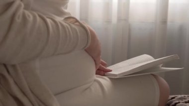 Otantik, tatlı, hamile bir kadın evde karnıyla mutlu hissediyor. Genç anne karnını okşuyor ve pencerenin yanındaki yatakta kitap okuyor. Doğum öncesi bakım, hamilelik, annelik, yaşam tarzı konsepti