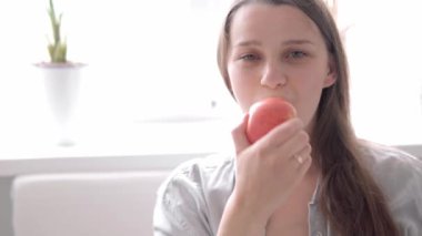 Otantik, tatlı, hamile bir kadın evde karnıyla mutlu hissediyor. Genç anne karnını okşuyor ve pencerenin yanındaki beyaz koltukta kırmızı yiyor. Doğum öncesi bakım, hamilelik, annelik, yaşam tarzı konsepti