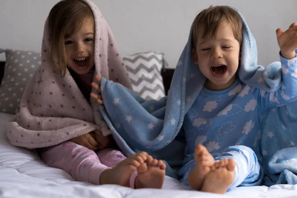 Otthon, kényelem, gyermekkor, ellátás, barátság - két vicces boldog autentikus baba kisgyermek testvér gyerekek ülnek a fehér ágyon nézd meg a kamerát csomagolva meleg kék puha takaró, hangulatos álmos hangulat beltérben — Stock Fotó
