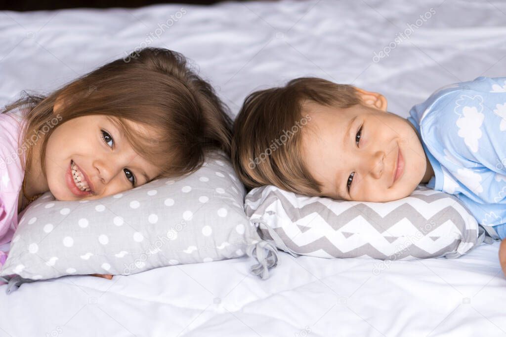 home, comfort, childhood, care, friendship, sweet dream- two smiling happy authentic toddler sibling kids children twins in pajamas lie rest sleep on bed pillows look at camera soft cozy sleepy mood