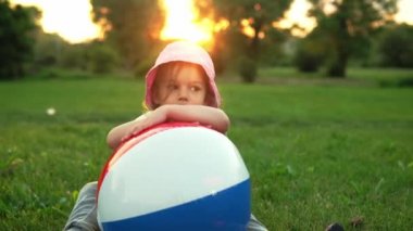 Yaz, tatil, doğa, mutlu çocukluk - pembe şapkalı, gülümseyen küçük bir anaokulu çocuğu kameraya poz veriyor ve gün batımında çayırda büyük bir şişme ile oynuyor.