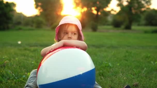 Lato, wakacje, przyroda, szczęśliwe dzieciństwo - jedno małe dziecko w przedszkolu uśmiechnięte dziecko dziewczyna w różowym kapeluszu siedzieć pozowanie patrząc na kamerę bawić się z dużym nadmuchiwane w parku na łące o zachodzie słońca świeże powietrze — Wideo stockowe