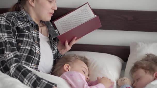 Authentic cute young woman or babysitter reads fairy tale from book to children before bedtime. Mom takes care of sleeping children. Two baby sleeping in white bed. Mothers day, motherhood, childhood — Stock Video