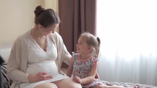 임신 한 여자는 집에서 아기를 돌보는 것을 행복하게 여긴다. 임신 한 젊은 어머니는 어린 딸을 배로 때리고 배에 입맞춤을 하였다. 출산 전 관리, 임신, 모성 개념. — 비디오