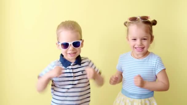 Familie, vriendschap, kindertijd, zomer. Twee blije Indrukwekkende glimlachende peuter kinderen broer of zus dans vieren spelen veel plezier lachen. Grappige kinderen in bril broer zus op geel kopiëren ruimte achtergrond — Stockvideo