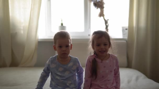 Dos niños sonrientes, niño y niña acostados en pijama sobre una cama blanca. Los hermanos gemelos pequeños se divierten, niños felices en cuarentena en casa. Amistad, familia, concepto educativo — Vídeos de Stock