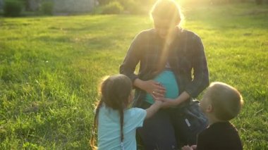 Yaz, doğa, mutlu aile, hamilelik - iki küçük çocuğu olan hamile kadın anne gün batımında parkta çimlerin üzerinde oturuyor. Çocuklar annelerinin karnını okşayıp eğleniyorlar, gülüyorlar birlikte vakit geçiriyorlar.
