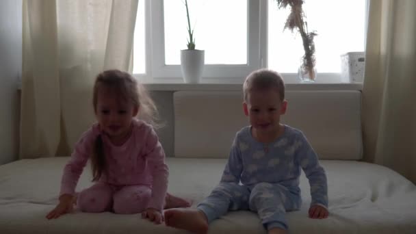 2人の笑顔の幼稚園、幼児の男の子と女の子が白いベッドの上のパジャマに腰を下ろしています。双子の兄弟は家で隔離されている楽しい幸せな子供を持っています。友情、家族、教育の概念 — ストック動画