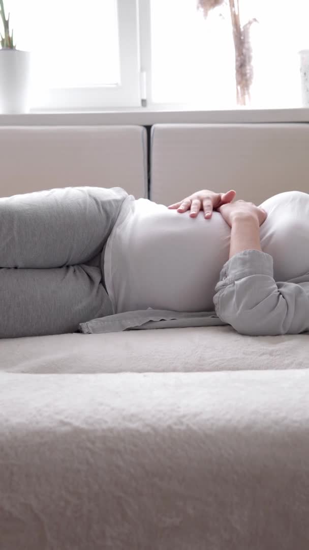 Yorgun, karnına dokunan hamile bir kadının güneşli bir günde evde yatıp dinlendiği dikey format videosu. hamilelik, annelik, insanlar, beklenti kavramı — Stok video