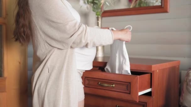 Милий щасливий вагітна жінка витягує дитячий одяг з комода, торкаючись її животик вдома. дбайлива мати гладить великий живіт руками на світлій кімнаті. вагітність, материнство, люди, концепція очікування — стокове відео